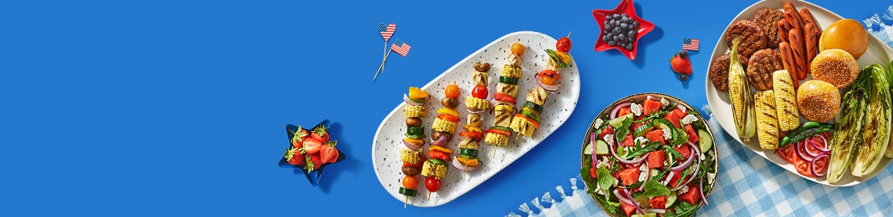 A festive summer cookout spread features grilled meats, vibrant vegetables, fresh fruit, and patriotic touches atop a blue checkered tablecloth.