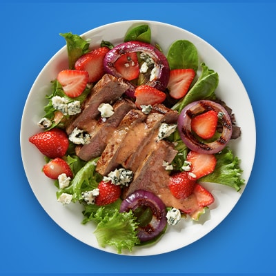 A vibrant salad composed of mixed greens, juicy strawberries, grilled red onion rings, crumbled blue cheese, and tender slices of medium-rare steak, all topped with a flavorful dressing.
