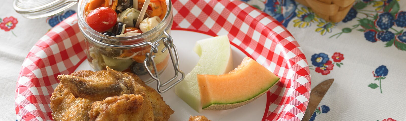 Red gingham paper plate loaded with fried chicken, fruit slices and a jar of pasta salad on a picnic blanket