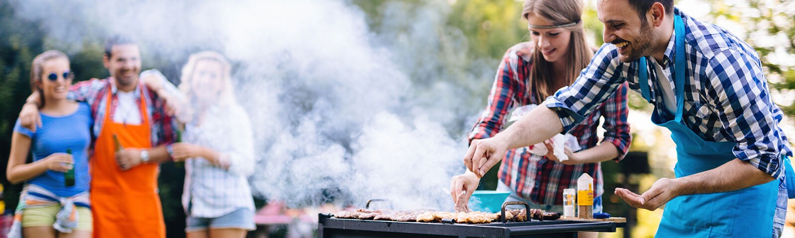 Outdoor grill with two people standing over it turning the hot dogs. In the background a group of friends watches on. Outdoor grill with two people standing over it turning the hot dogs. In the background a group of friends watches on.
